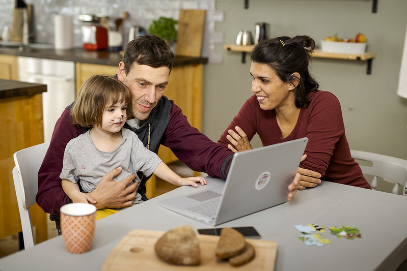 Ein mittelaltes Elternpärchen sitzt am Tisch. Der Vater hat seine Tochter auf dem Schoß.