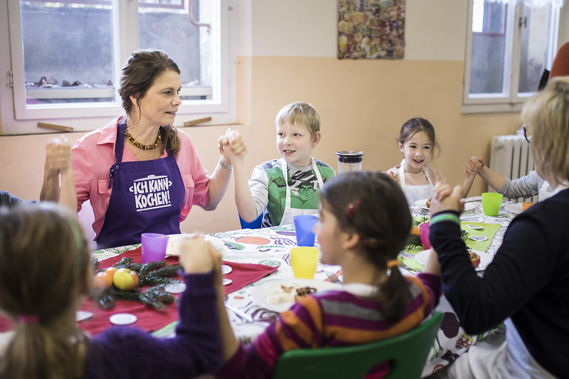 Sarah Wiener sitzt am bunt gedeckten Tisch mit Kindern im Alter zw. 3 und 10 Jahren und hält Händchen.